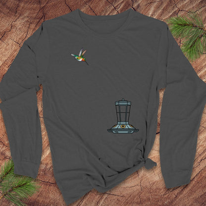 A dark gray long-sleeve t-shirt with a hummingbird and bird feeder graphic, laid flat on a rustic wood background with pine needles.