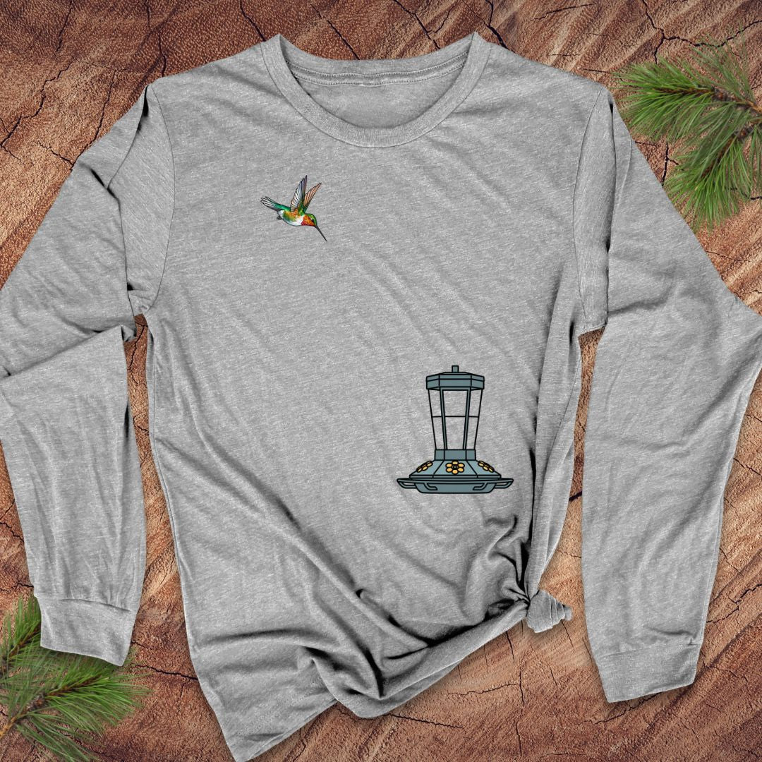 A heather gray long-sleeve t-shirt with a hummingbird and bird feeder graphic, laid flat on a rustic wood background with pine needles.