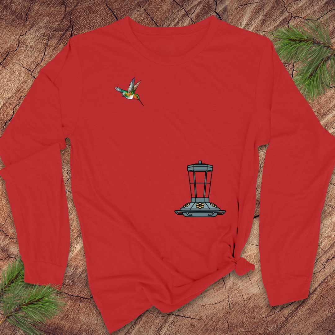 A red long-sleeve t-shirt with a hummingbird and bird feeder graphic, laid flat on a rustic wood background with pine needles.