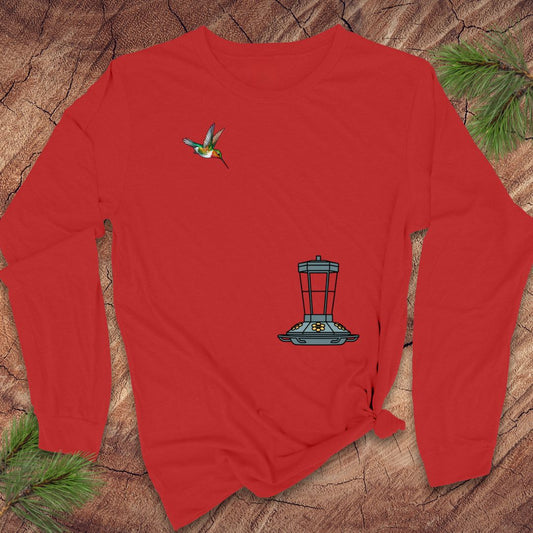 A red long-sleeve t-shirt with a hummingbird and bird feeder graphic, laid flat on a rustic wood background with pine needles.