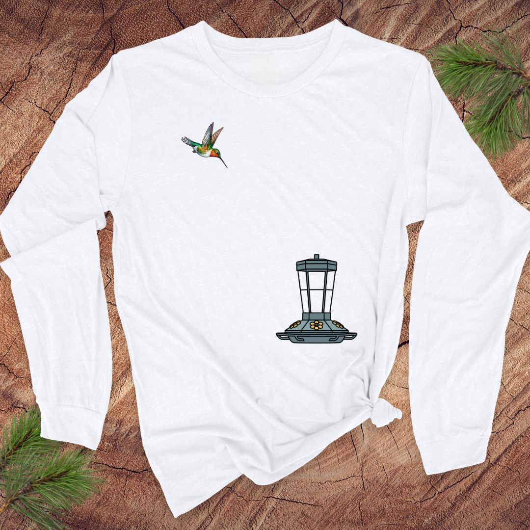 A white long-sleeve t-shirt with a hummingbird and bird feeder graphic, laid flat on a rustic wood background with pine needles.