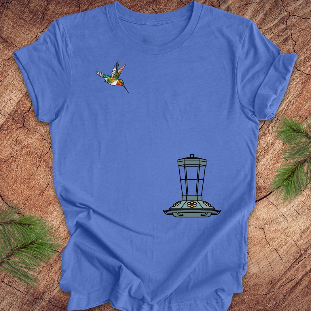 A blue short-sleeve crewneck t-shirt laid flat on a rustic wood background with pine needles in the corners. The shirt features a graphic of a hummingbird flying near a bird feeder.