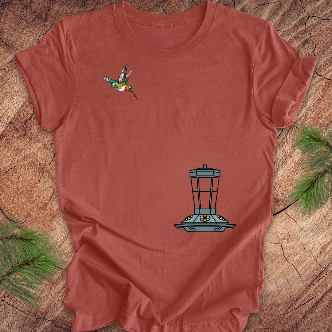 A terracotta-colored short-sleeve t-shirt laid flat on a rustic wood slice background, decorated with pine needles. The shirt features a graphic of a hummingbird on the left chest and a blue bird feeder on the lower right chest.