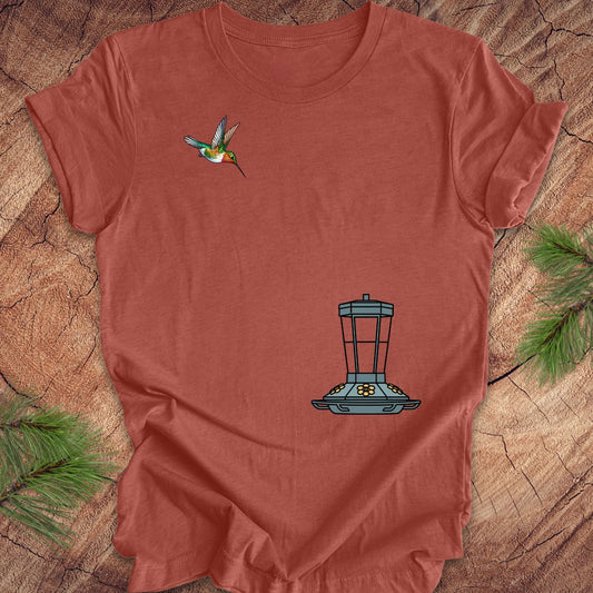 A terracotta-colored short-sleeve t-shirt laid flat on a rustic wood slice background, decorated with pine needles. The shirt features a graphic of a hummingbird on the left chest and a blue bird feeder on the lower right chest.