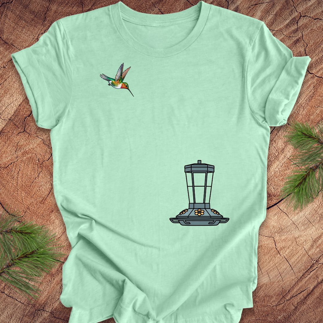 A mint green short-sleeve t-shirt laid flat on a rustic wood surface, featuring a small hummingbird graphic on the upper left chest and a black-and-gray bird feeder graphic on the lower right chest. Pine branches are placed around the edges of the frame.
