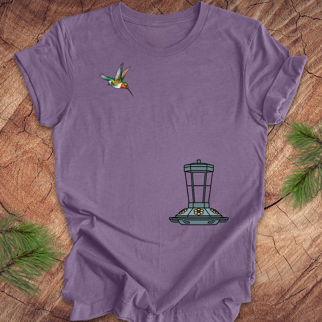 A purple short-sleeve t-shirt laid flat on a rustic wood surface, featuring a small hummingbird graphic on the upper left chest and a black-and-gray bird feeder graphic on the lower right chest. Pine branches are placed around the edges of the frame.