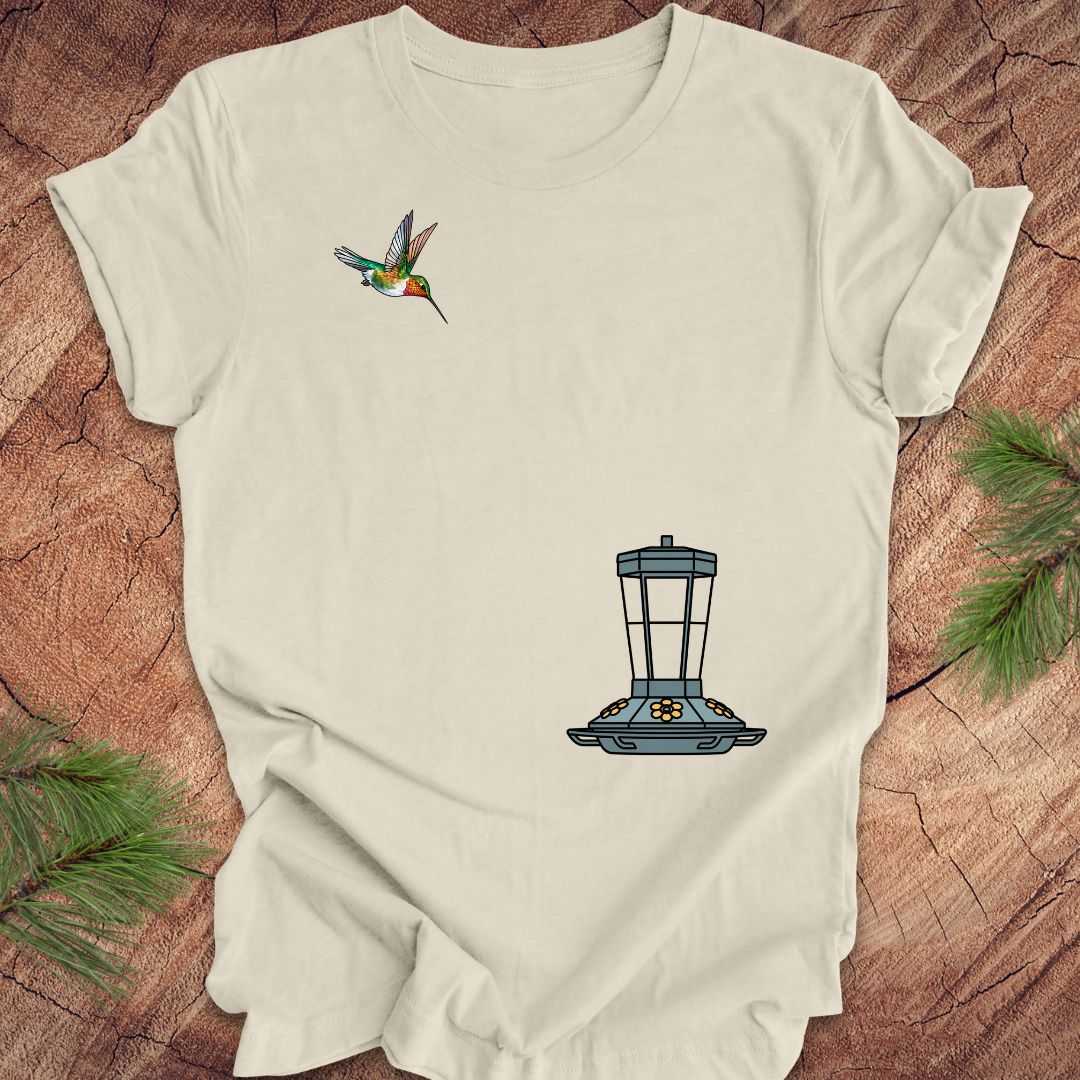 An off-white short-sleeve t-shirt laid flat on a rustic wood background with pine branches. The shirt features a graphic of a hummingbird on the left chest and a blue bird feeder on the lower right chest.