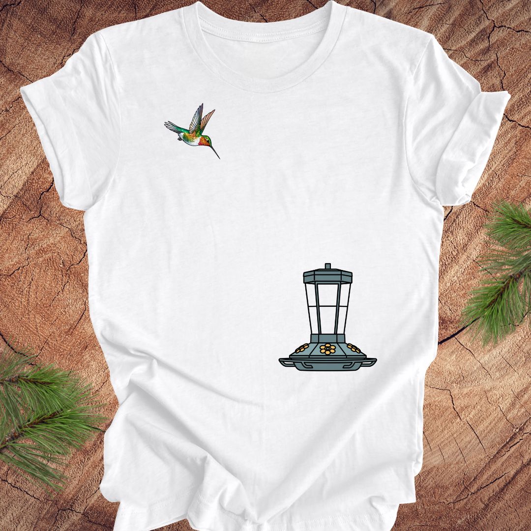 A white short-sleeve t-shirt laid flat on a rustic wood surface, featuring a small hummingbird graphic on the upper left chest and a larger bird feeder graphic on the lower right chest. Pine branches are visible at the corners of the frame.