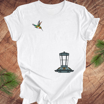 A white short-sleeve t-shirt laid flat on a rustic wood surface, featuring a small hummingbird graphic on the upper left chest and a larger bird feeder graphic on the lower right chest. Pine branches are visible at the corners of the frame.