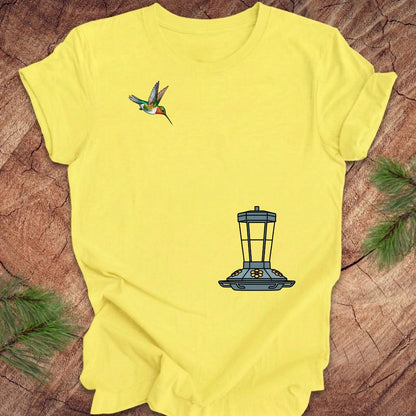 A bright yellow short-sleeve t-shirt laid flat on a textured wood surface, decorated with pine needles in the corners. The shirt features a graphic of a hummingbird on the left chest and a blue bird feeder on the lower right front.
