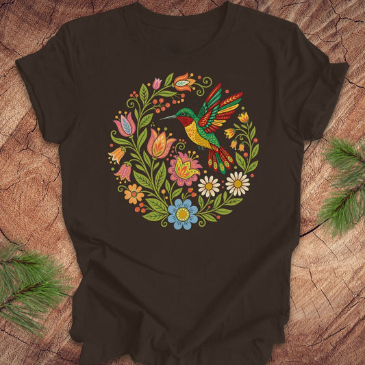 A dark brown short-sleeve t-shirt with a colorful embroidered-style circular graphic of a hummingbird surrounded by flowers, laid flat on a wood surface with pine branches in the corners.