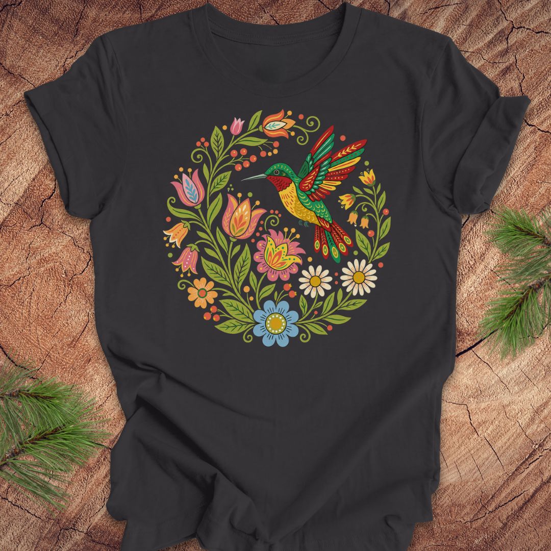 A black short-sleeve crewneck t-shirt featuring a colorful circular graphic of a hummingbird surrounded by flowers, laid flat on a rustic wood background with pine branches in the corners.