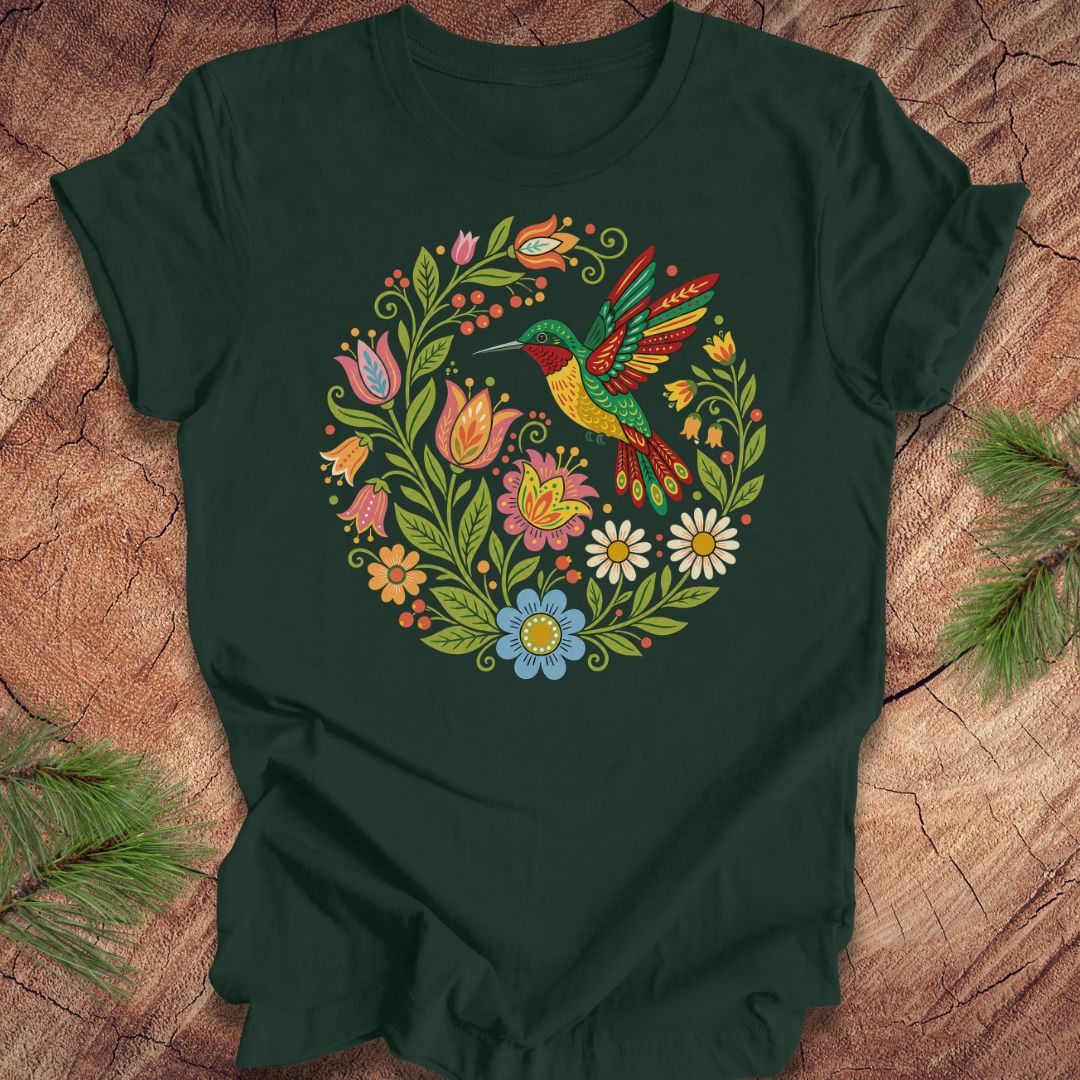 A dark green short-sleeve t-shirt laid flat on a rustic wood surface, featuring a colorful circular graphic of a hummingbird surrounded by flowers. Pine branches are placed in the corners of the frame.