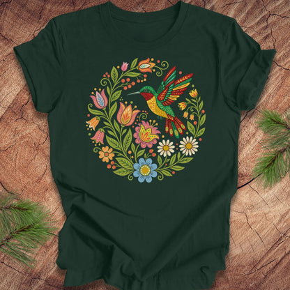 A dark green short-sleeve t-shirt laid flat on a rustic wood surface, featuring a colorful circular graphic of a hummingbird surrounded by flowers. Pine branches are placed in the corners of the frame.