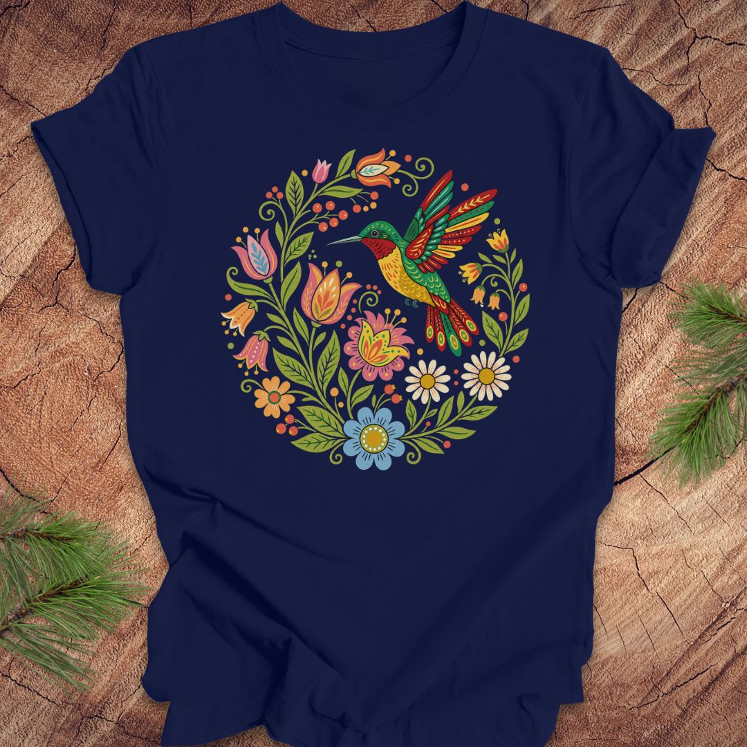 A navy blue short-sleeve t-shirt laid flat on a rustic wood surface, featuring a colorful circular graphic of a hummingbird surrounded by flowers. Green pine branches are placed in the corners of the frame.