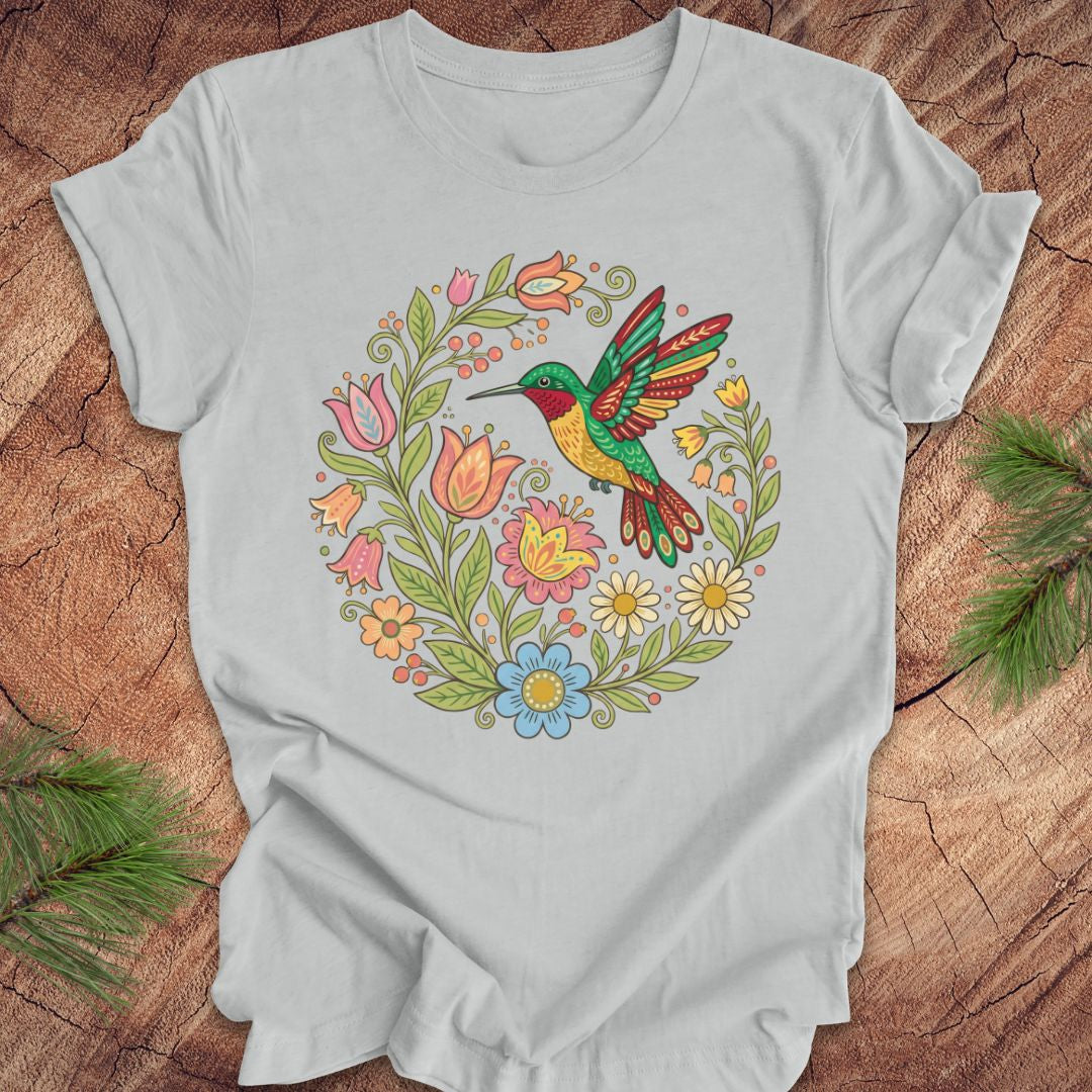 Silver t-shirt with a colorful hummingbird and floral design on a wooden surface.