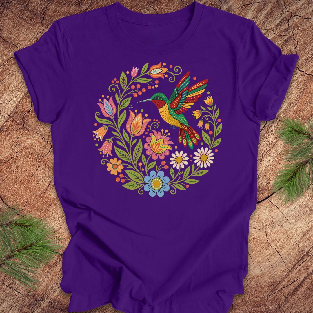 A purple short-sleeve crewneck t-shirt featuring a colorful circular graphic of a hummingbird surrounded by flowers, laid flat on a rustic wood background with pine branches in the corners.