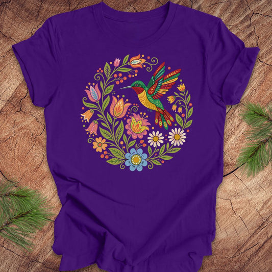 A purple short-sleeve crewneck t-shirt featuring a colorful circular graphic of a hummingbird surrounded by flowers, laid flat on a rustic wood background with pine branches in the corners.
