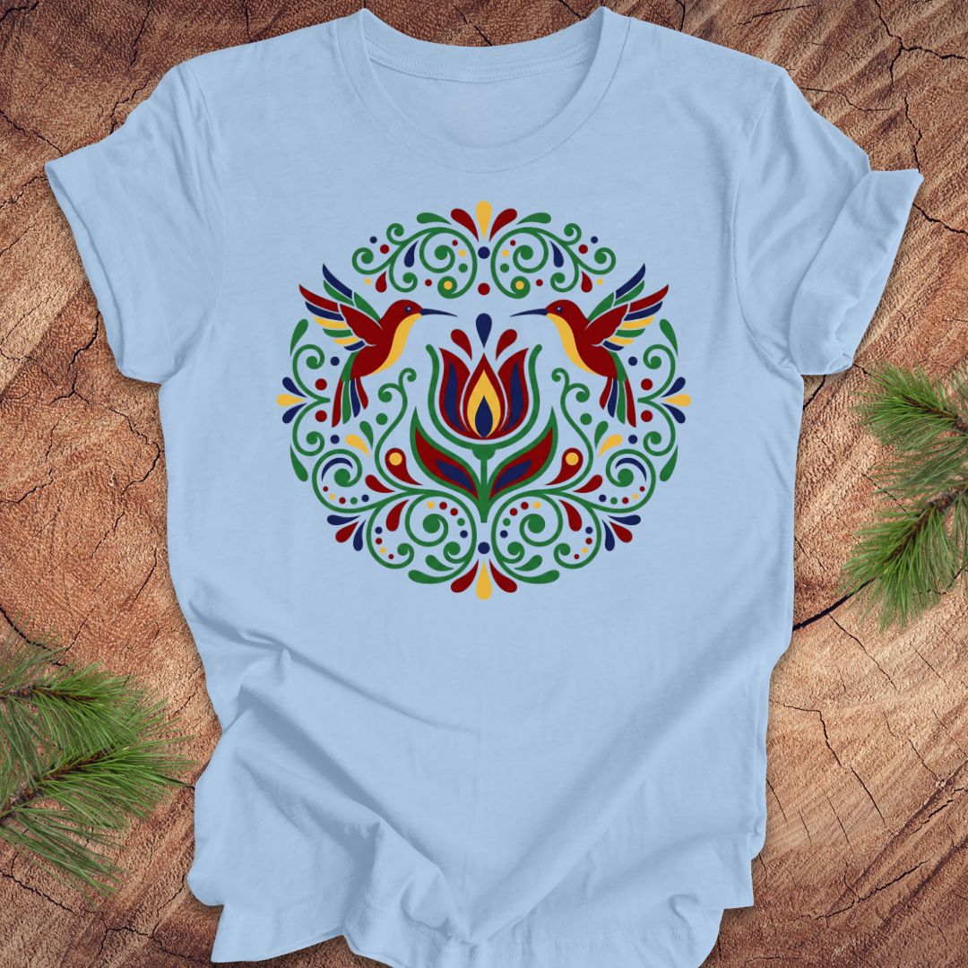 Light blue t-shirt with colorful floral design and hummingbirds on a wooden surface