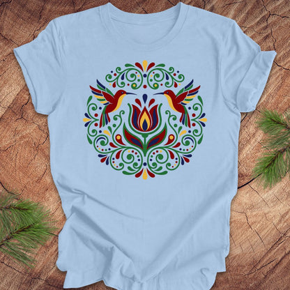 Light blue t-shirt with colorful floral design and hummingbirds on a wooden surface