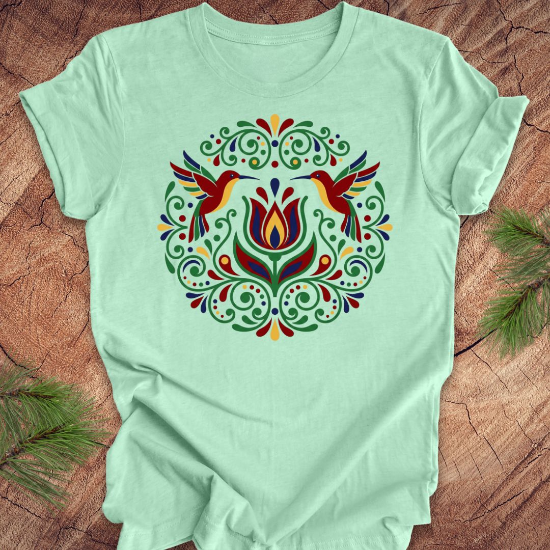 Light green t-shirt with colorful floral design and hummingbirds on a wooden surface