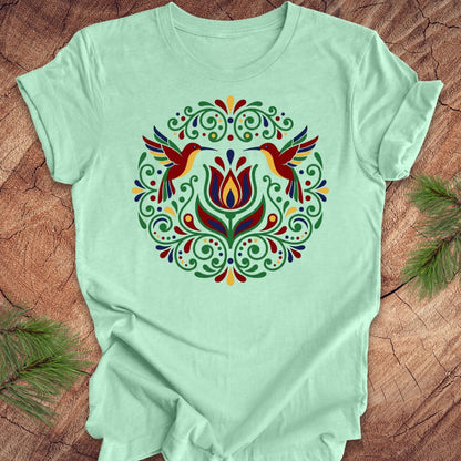 Light green t-shirt with colorful floral design and hummingbirds on a wooden surface