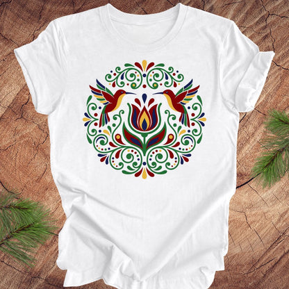 White t-shirt with colorful floral design and hummingbirds on a wooden surface