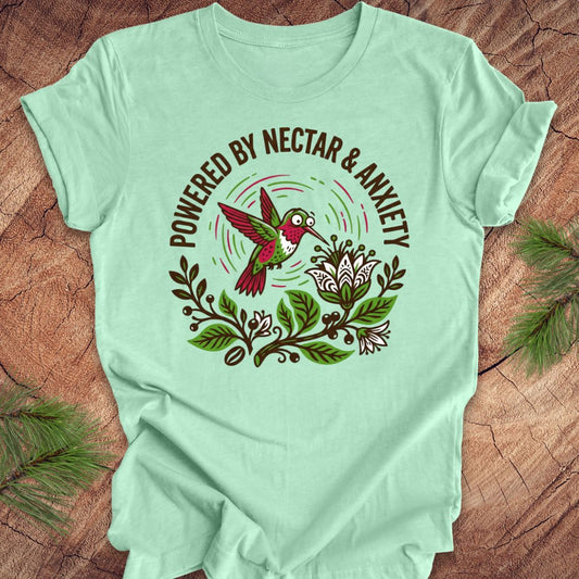 Mint green t-shirt with a colorful hummingbird graphic and text 'Powered by Nectar & Anxiety' on a wooden surface.