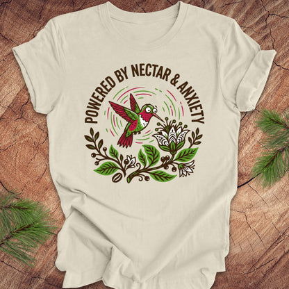 Beige t-shirt with a colorful hummingbird graphic and text 'Powered by Nectar & Anxiety' on a wooden surface.
