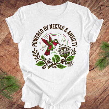 White t-shirt with a colorful hummingbird graphic and text 'Powered by Nectar & Anxiety' on a wooden surface.