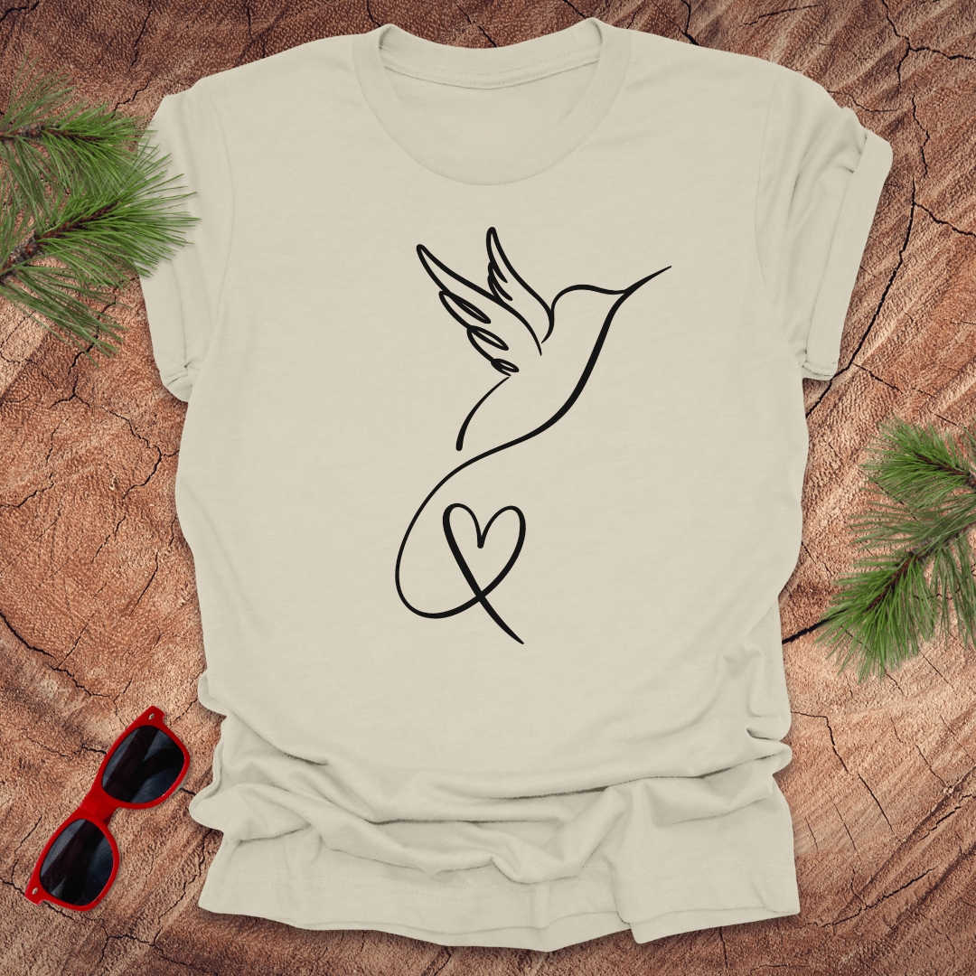 Beige t-shirt with hummingbird and heart design on a wooden surface with sunglasses and greenery.