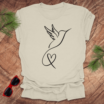Beige t-shirt with hummingbird and heart design on a wooden surface with sunglasses and greenery.
