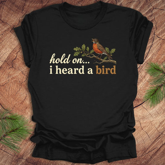 Black t-shirt with a bird graphic and text 'hold on...i heard a bird' on a wooden background