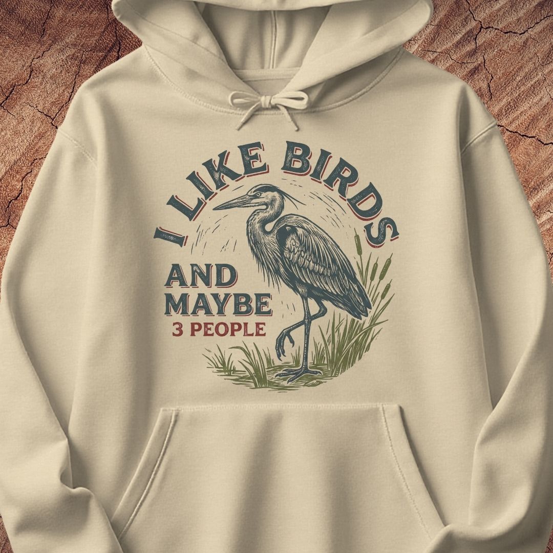 An off-white hooded sweatshirt with a graphic print of a heron and the text 'I LIKE BIRDS AND MAYBE 3 PEOPLE', displayed against a textured wood background.