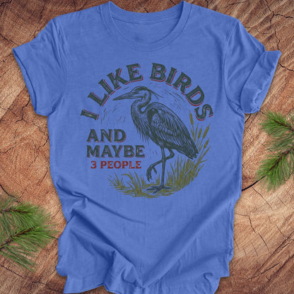 Blue t-shirt with heron graphic and text, 'i like birds and maybe 3 people' on a wooden surface