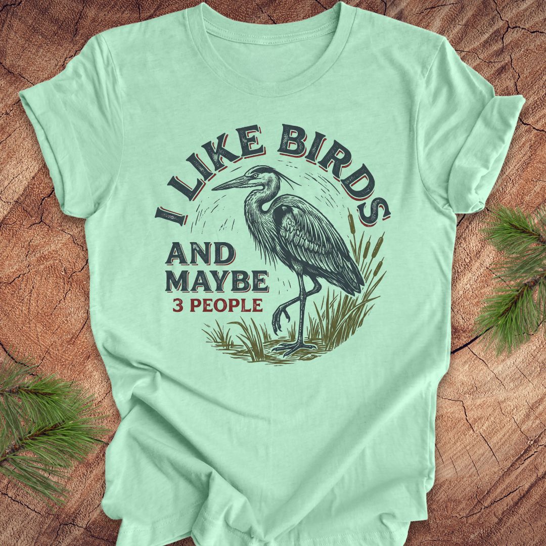 Mint green t-shirt with heron graphic and text, 'i like birds and maybe 3 people' on a wooden surface