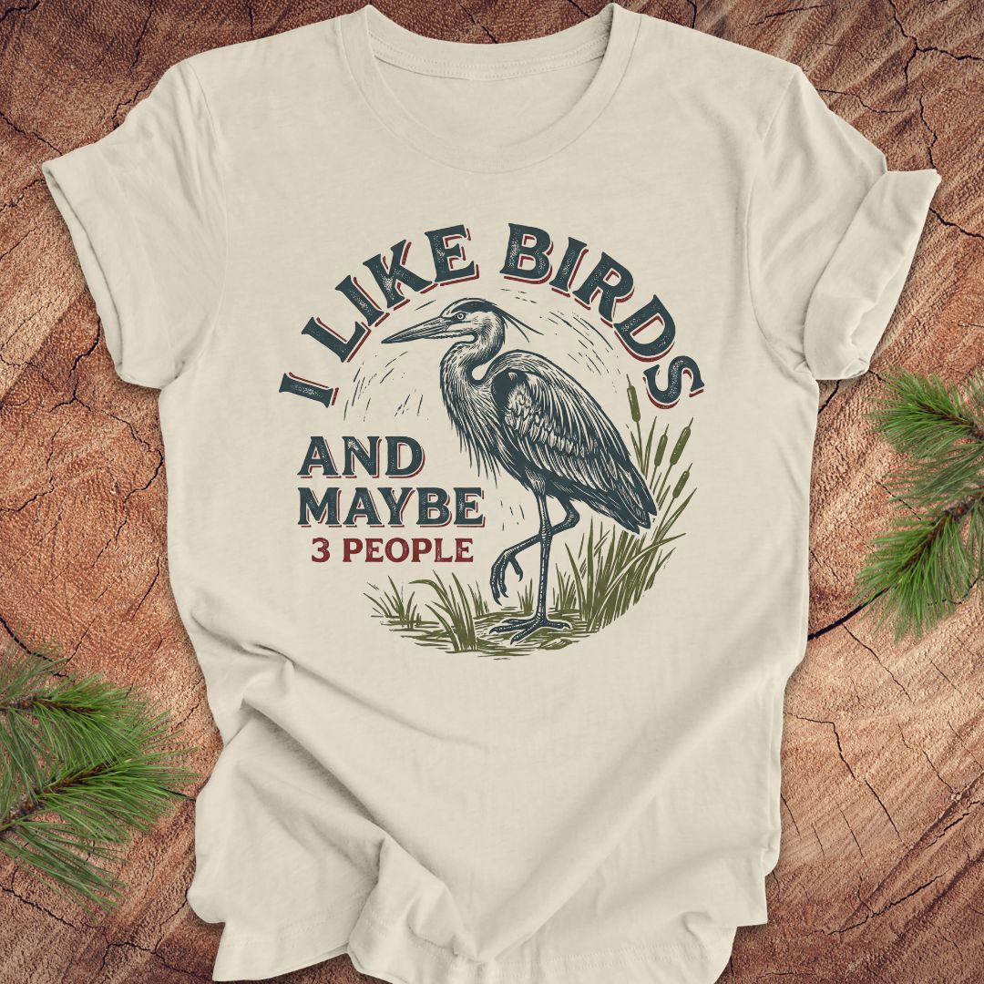 Beige t-shirt with heron graphic and text, 'i like birds and maybe 3 people' on a wooden surface
