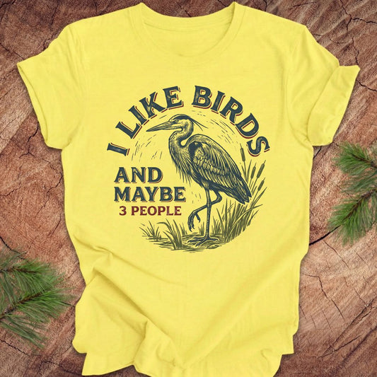 Yellow t-shirt with heron graphic and text, 'i like birds and maybe 3 people' on a wooden surface