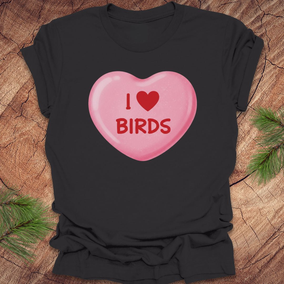 Black t-shirt with a pink heart-shaped candy design with text, 'i love birds' on a wooden surface with greenery.
