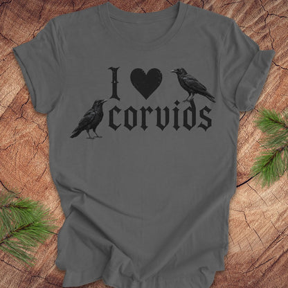 A dark gray short-sleeve t-shirt with a black graphic print reading 'I [heart] corvids' with two black raven illustrations, laid flat on a rustic wood background with pine branches in the corners.