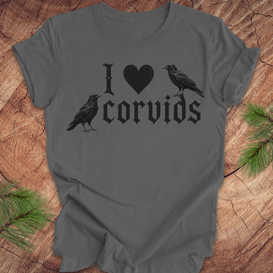 A dark gray short-sleeve t-shirt with a black graphic print reading 'I [heart] corvids' with two black raven illustrations, laid flat on a rustic wood background with pine branches in the corners.