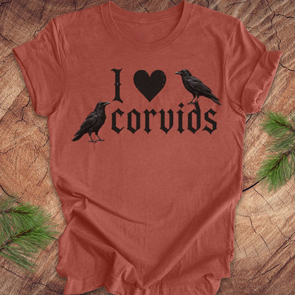 A terracotta-colored t-shirt laid flat on a rustic wood background with pine branches in the corners. The shirt features a black graphic print that reads 'I' followed by a heart, then 'corvids', with two black raven illustrations integrated into the design.