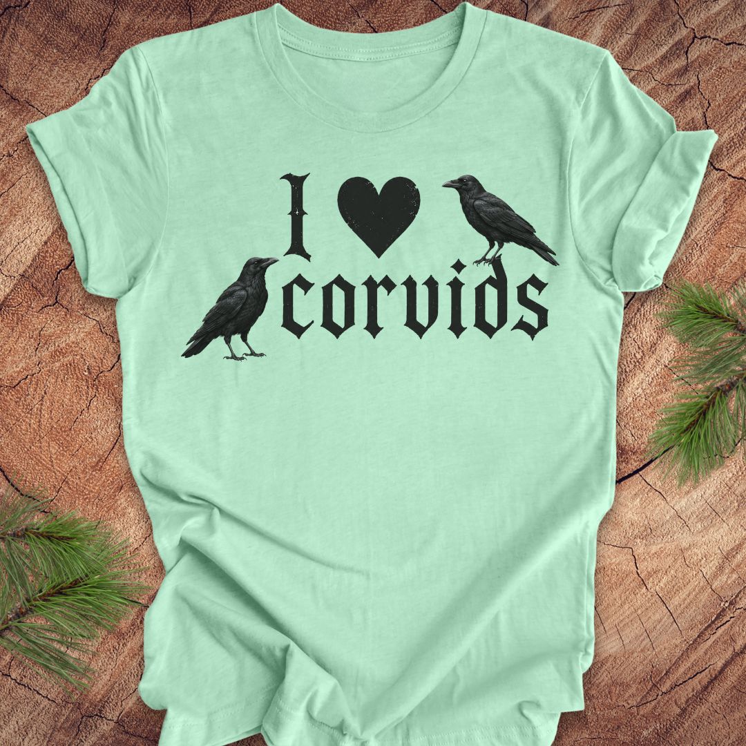 A mint green short-sleeve t-shirt with a black graphic reading 'I [heart] corvids' with two crow silhouettes, laid flat on a rustic wood surface with pine branches in the corners.