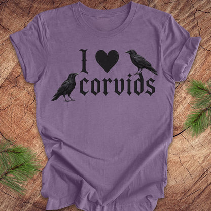 A purple short-sleeve t-shirt with a black graphic reading 'I [heart] corvids' with two black raven illustrations, laid flat on a rustic wood background with pine branches in the corners.