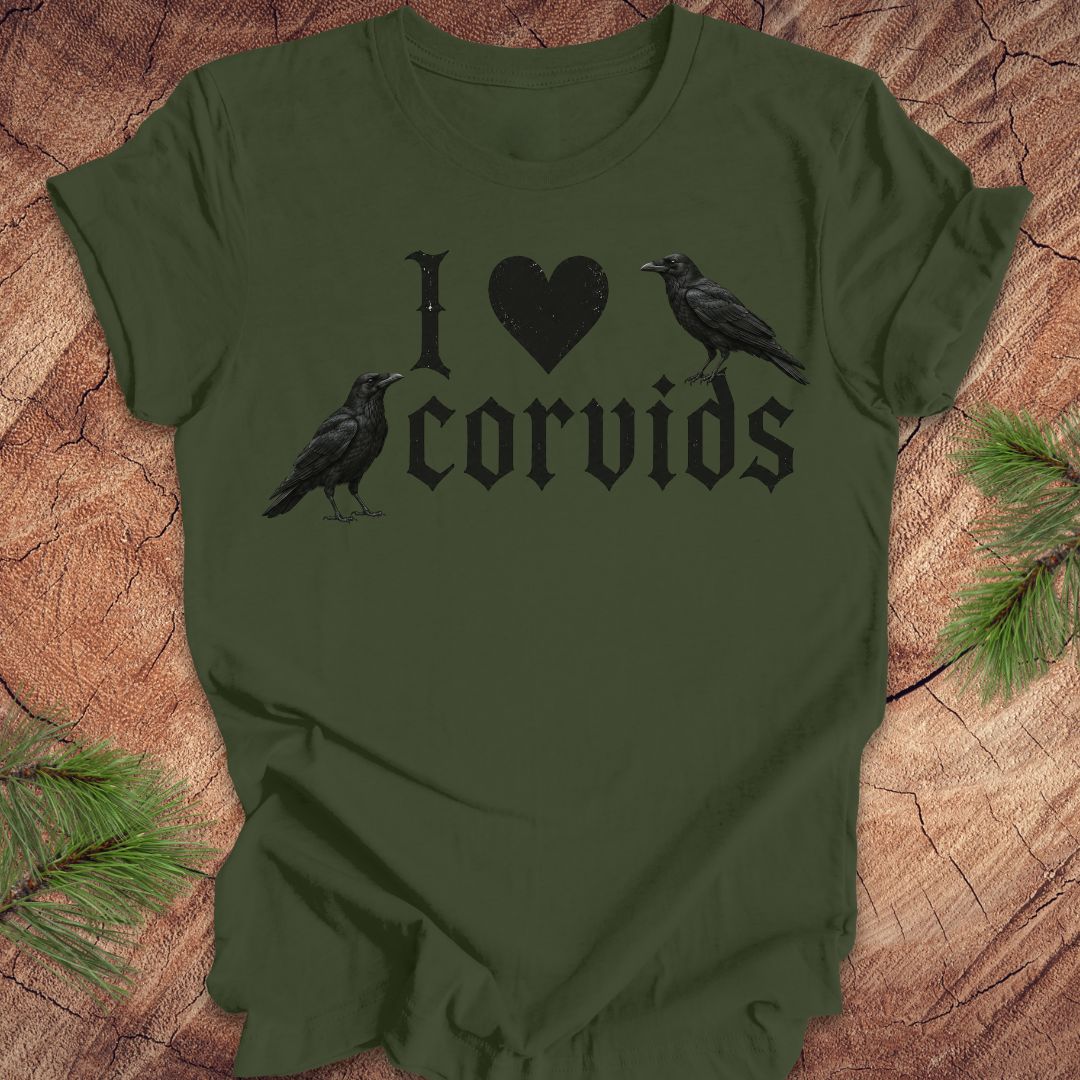 An olive green short-sleeve t-shirt with a black graphic reading 'I [heart] corvids' with two black raven illustrations, laid flat on a rustic wood background with pine branches in the corners.