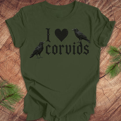 An olive green short-sleeve t-shirt with a black graphic reading 'I [heart] corvids' with two black raven illustrations, laid flat on a rustic wood background with pine branches in the corners.