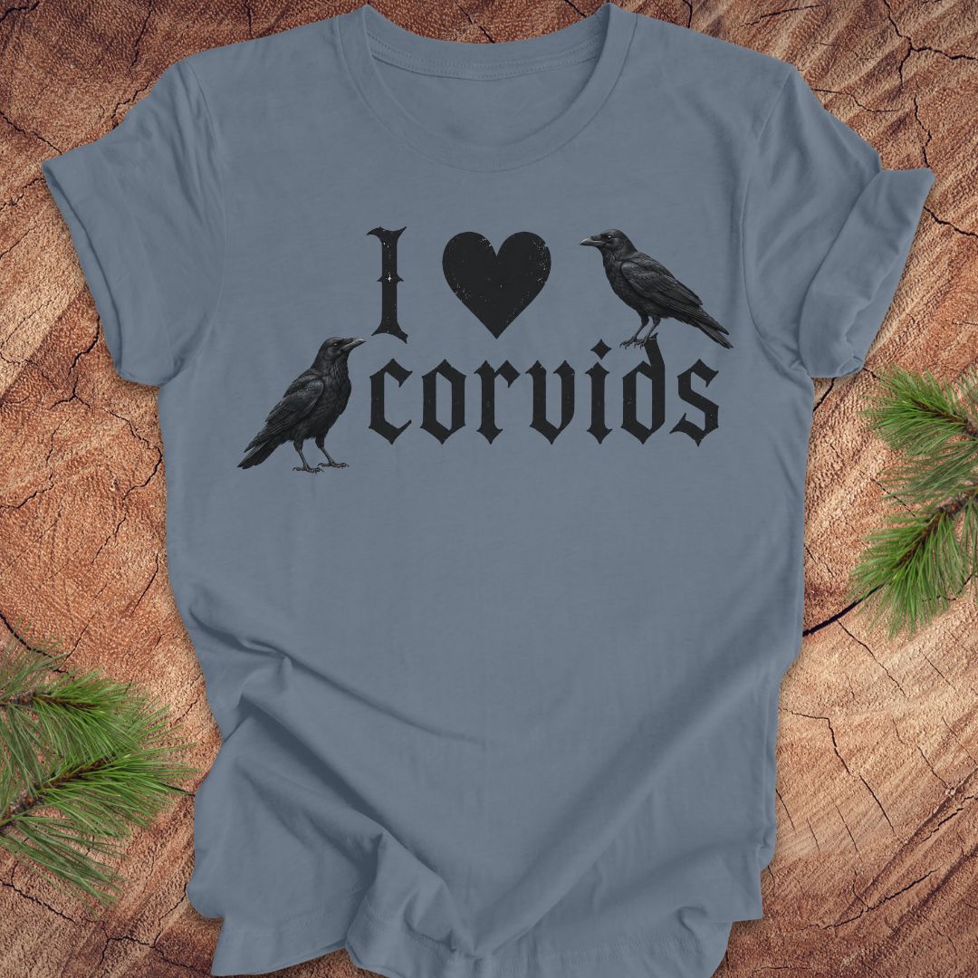 A slate blue short-sleeve t-shirt with a black graphic print reading 'I [heart] corvids' with two black raven illustrations, displayed flat on a rustic wood background with pine needles.