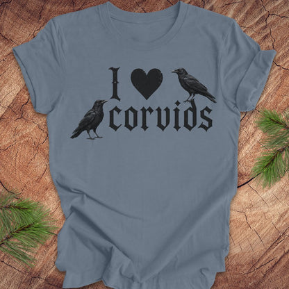 A slate blue short-sleeve t-shirt with a black graphic print reading 'I [heart] corvids' with two black raven illustrations, displayed flat on a rustic wood background with pine needles.