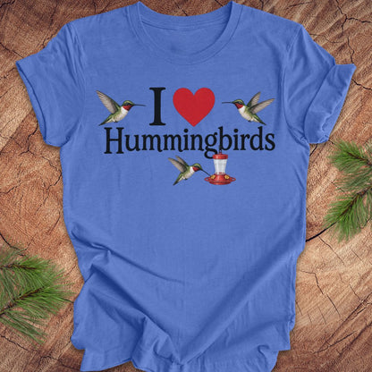 A blue short-sleeve crewneck t-shirt laid flat on a rustic wood background with pine branches in the corners. The shirt features a graphic print that reads 'I [red heart] Hummingbirds' with illustrations of hummingbirds and a red-and-white hummingbird feeder.