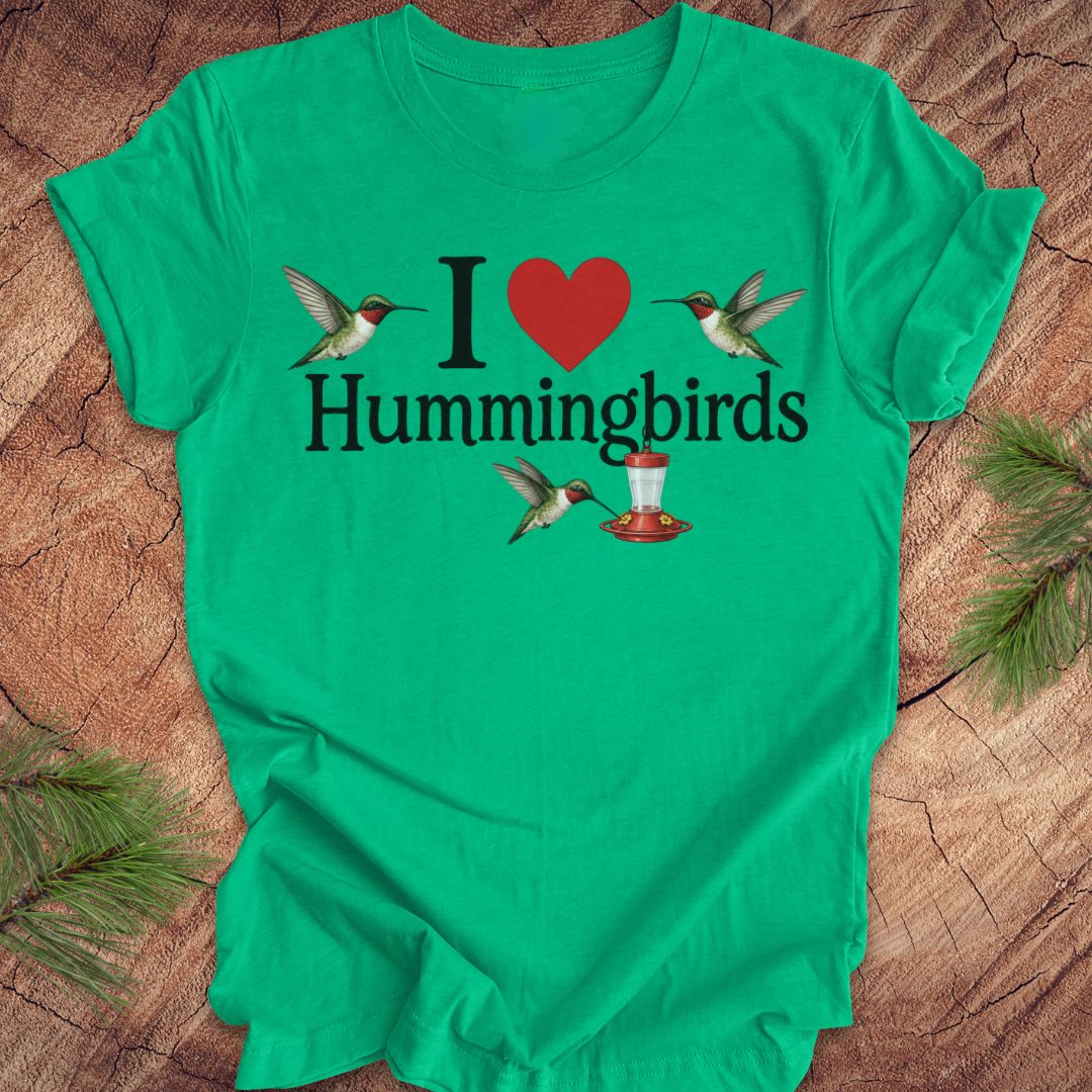 A bright green short-sleeve t-shirt laid flat on a rustic wood surface, featuring a graphic that reads 'I [red heart] Hummingbirds' with illustrated hummingbirds and a red-and-white hummingbird feeder.